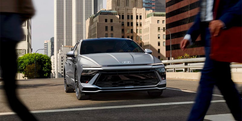 Silver sedan driving on a city street past a pedestrian.