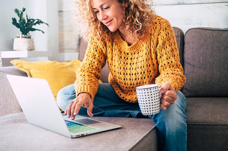 A woman sits on a sofa using a laptop while holding a mug